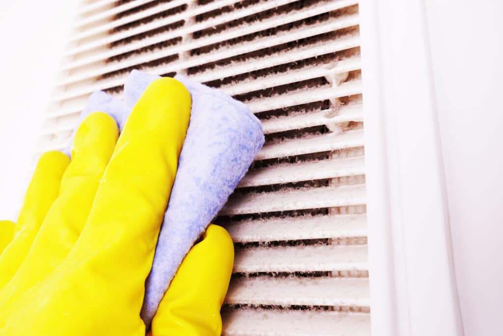 person cleaning a dusty air duct vent with a towel while wearing yellow gloves
